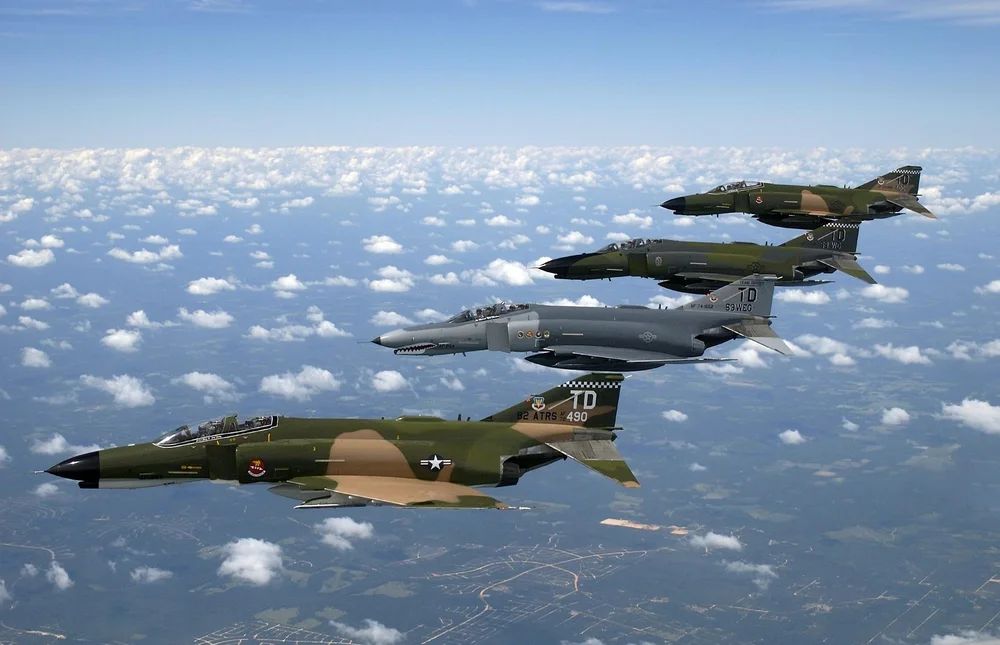 Four real F-4 aircraft in formation above a cloud-dotted landscape